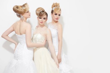 Parlak makyajlı ve zarif saç stili olan beyaz gelinlikli üç güzel sarışın kız. Düğün makyajı ve saç stili. Açık gri arkaplanda fotokopi alanı olan bir stüdyo portresi.