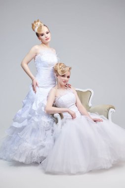 Öncü saç stili ve makyajlı, beyaz gelinlikler içinde poz veren iki sarışın kadının tam boy portresi. Stüdyo görüntüsü gri arka planda. Düğün Modası. 