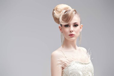 Moda ve düğün tarzı. Beyaz gelinlik içinde, abartılı saçları ve makyajı olan güzel sarışın bir kızın portresi. Açık gri arkaplanda fotokopi alanı olan bir stüdyo görüntüsü.