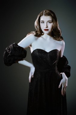 Şık bir akşam makyajı ve saç stili. Stüdyo arka planında lüks siyah elbiseli çekici bir genç kadının portresi. Eski Hollywood tarzı.