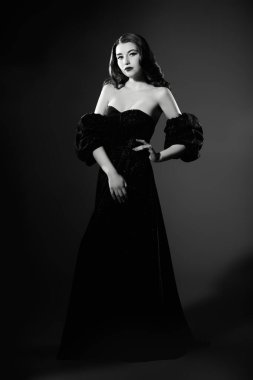 Siyah dalgalı saçlı, zarif makyajlı, siyah dekolteli bir elbiseyle siyah bir stüdyoda poz veren siyah ve beyaz bir kadının tam boy portresi. Eski Hollywood tarzı.