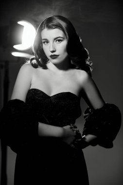 Siyah dalgalı saçlı, zarif makyajlı siyah dekolteli siyah bir elbisenin içinde duran güzel bir bayanın siyah beyaz portresi. Eski Hollywood tarzı.