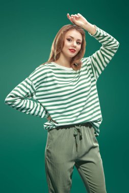 Yeşil pantolonlu, yeşil çizgili uzun kollu, eğlenceli pozlar veren mutlu bir kız. Yeşil arka planda stüdyo portresi. Aziz Patrick Günü kutlaması.