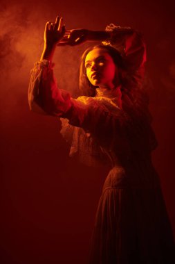 Uzun beyaz dantelli bir elbise içindeki güzel romantik bir kızın portresi, puslu bir şekilde gizlenmiş bir ışık demetine bakıyor. Sanat portresi. Stüdyo çekimi koyu kırmızı arka planda.