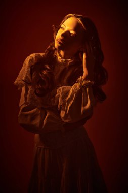 Uzun beyaz dantelli elbisesi olan, sıcak bir ışık huzmesiyle aydınlatılmış güzel bir kız. Sanat fantezisi. Stüdyo çekimi koyu kırmızı arka planda. Klasik stil.