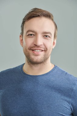 Mavi tişörtlü, kameraya gülümseyen orta yaşlı bir adamın portresi. İnsanlar, duygular. Erkek sağlığı ve güzelliği. Gri arkaplanda stüdyo portresi. 