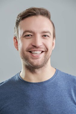 Mavi tişörtlü, kameraya gülümseyen orta yaşlı bir adamın pozu. İnsanlar, duygular. Erkek sağlığı ve güzelliği. Gri arkaplanda stüdyo portresi. 