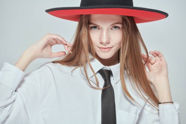 Moda ve tarz. Klasik tarz kıyafetlerle stüdyoda poz veren güzel bir manken kızın portresi. Beyaz bir bluz, siyah kravat ve geniş kenarlı kırmızı ve siyah şapka. Beyaz arkaplan.