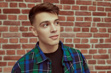 Kareli gömlekli yakışıklı, sakin bir delikanlının tuğla bir duvara karşı fotoğrafı. Gençlik modası. İnsanlar ve duygular. Stüdyo resmi.