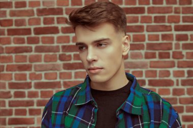 Kareli gömlekli, tuğla bir duvara yaslanmış yakışıklı, sakin bir genç. Gençlik modası. İnsanlar ve duygular. Stüdyo resmi.