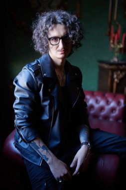 Esmer, kıvırcık saçlı, gözlüklü, siyah deri ceketli, vücudunda dövmeler olan ve klasik bir iç mimarisi olan görkemli bir adamın portresi. Gayri resmi hipster.