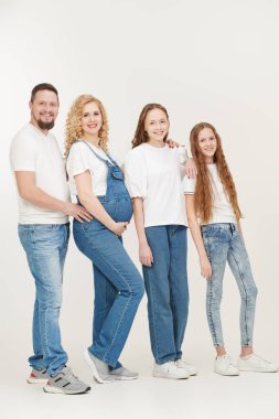 Mutlu ebeveynler ve sevgili kızları beyaz tişörtler ve kot pantolonlar içinde beyaz stüdyo arka planında birlikte poz veriyorlar. Mutlu ve dost canlısı bir aile. Güzel hamile anne..