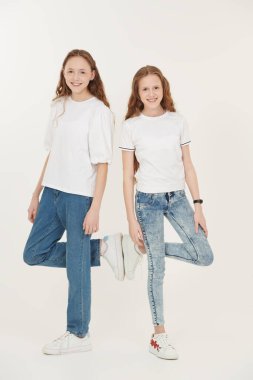 Beyaz tişörtlü iki mutlu kız kardeş yan yana durur ve gülümser. Beyaz arka planda stüdyo boy portresi. Mutlu bir aile ve çocukluk. Çocuklar ve gençler için gündelik moda.