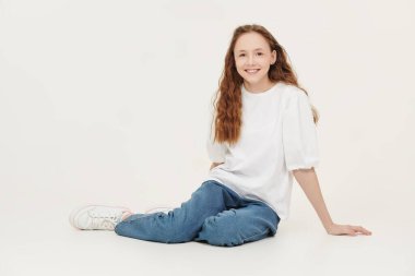 Uzun dalgalı saçlı, beyaz bluzlu ve kameraya gülümseyen kot pantolonlu güzel bir genç kızın tam boy portresi. Beyaz stüdyo geçmişi.