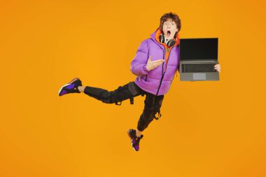 Eğitim. Şık parlak elbiseli genç bir çocuk havada uçuyor, önünde dizüstü bilgisayar tutuyor ve çok şaşırıyor. Stüdyo sarı arka plan. Modern gençlerin yaşam tarzı..