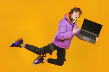 Şık parlak elbiseli genç bir çocuk havada uçuyor, önünde dizüstü bilgisayar tutuyor ve çok şaşırıyor. Stüdyo sarı arka plan. Modern gençlerin yaşam tarzı. Eğitim.