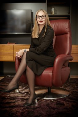 Modern bir ofiste pahalı bir deri koltukta oturan ve gülümseyen zarif, orta yaşlı bir kadın. Zarif siyah elbise ve gözlüklü başarılı bir iş kadını..