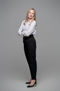 Orta yaşlı mutlu bir iş kadınının tam boy portresi. Klasik resmi kıyafet ve gözlüklerle kameraya bakıp gülümsüyor. Gri stüdyo geçmişi.