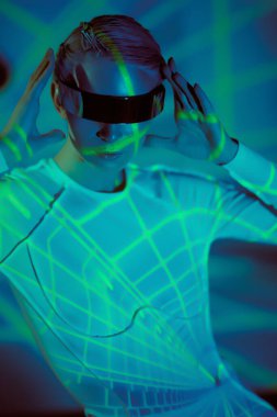 Fütürist gözlüklü ve tulumlu mükemmel siber kız yeşil neon ışıkların arasında duruyor. Siber uzay. Geleceğin dijital teknolojileri. VR oyunları.