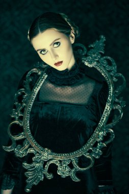 Zarif siyah kadife elbise ve dantel eldivenli dramatik esmer bir kızın portresi, gecenin karanlığında ayna çerçeveli esmer bir arka planda duruyor. Gotik roman, mistisizm..