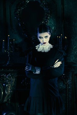 Addams Ailesi. Beyaz yakalı, düzgün siyah elbiseli esmer bir kız kasvetli gotik bir kalede duruyor ve kameraya pek dostça görünmüyor. Gotik kara komedi.