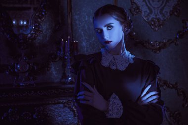 Beyaz yakalı, düzgün siyah elbiseli esmer bir kadın kasvetli gotik bir şatoda gece ayakta duruyor ve kameraya pek dostça görünmüyor. Gotik kara komedi. Addams Ailesi. 