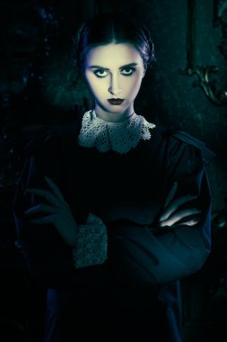 Beyaz yakalı, düzgün siyah elbiseli esmer bir kadın kasvetli gotik bir şatoda gece ayakta duruyor ve kameraya pek dostça görünmüyor. Gotik kara komedi. Addams Ailesi. 