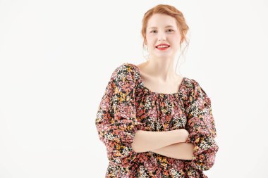 Çiçekli bir elbisenin içinde beyaz bir duvara karşı duran zarif makyajlı güzel bir kızıl kızın stüdyo fotoğrafı. Romantik kadın tarzı. Bahar ve yaz modası, güzellik. Boşluğu kopyala.