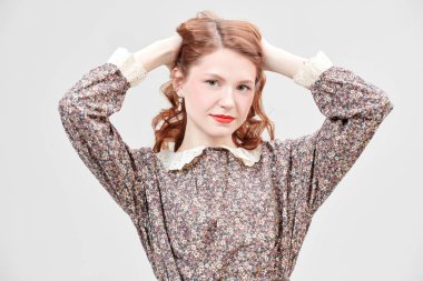 Çiçekli bir elbisenin içinde beyaz bir duvara karşı duran zarif makyajlı güzel bir kızıl kızın stüdyo fotoğrafı. Romantik kadın tarzı. Bahar ve yaz modası, güzellik.