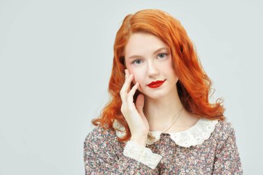 Çiçekli bir elbisenin içinde beyaz bir duvara karşı duran zarif makyajlı güzel bir kızıl kızın stüdyo fotoğrafı. Bahar ve yaz modası, güzellik. Romantik kadınsı stil. 