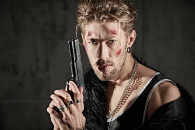 Yüzünde morluklar ve yaralar olan, beton duvarlı karanlık bir odada elinde silahla kameraya saldırganca bakan sert bir erkek haydut vuruldu. Aksiyon filmi kahramanı. Kötü adam konsepti.