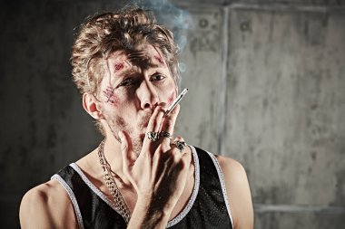 Kötü adam konsepti. Yüzünde morluklar ve yaralar olan, beton duvarlı karanlık bir odada sigara içen sert bir erkek haydutun fotoğrafı. Aksiyon filmi kahramanı.