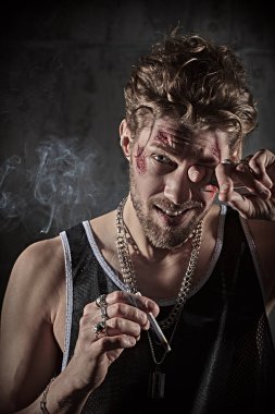 Yüzünde morluklar ve yaralar olan, beton duvarlı karanlık bir odada sigara içen sert bir erkek haydutun fotoğrafı. Kötü adam konsepti. Aksiyon filmi kahramanı.
