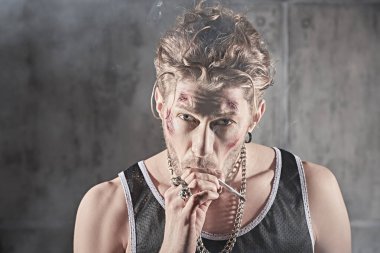 Yüzünde morluklar ve yaralar olan, beton duvarlı karanlık bir odada sigara içen sert bir erkek haydutun fotoğrafı. Kötü adam konsepti. Aksiyon filmi kahramanı.