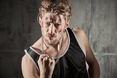 Yüzünde morluklar ve yaralar olan, beton duvarlarla karanlık bir odada duran ve saldırganca bakan sert bir erkek haydudun fotoğrafı. Kötü adam konsepti. Aksiyon filmi kahramanı.