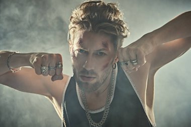 Yüzünde morluklar ve yaralar olan, beton duvarlarla karanlık bir odada duran ve saldırganca bakan sert bir erkek haydudun fotoğrafı. Kötü adam konsepti. Aksiyon filmi kahramanı.