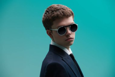 Klasik beyaz bir gömlek, şık bir takım elbise ve mavi bir arka planda şık yuvarlak güneş gözlüğü takan yakışıklı, bakımlı genç bir adam. Erkek modası, klasik tarz ve güzellik. Gözlük stili.