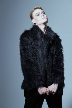 Kısa saç kesimi ve siyah dumanlı gözleriyle stüdyoda siyah kürklü poz veren şık bir gencin portresi. Makyaj, saç stili, makyaj malzemeleri. Moda ve erkek güzelliği. 