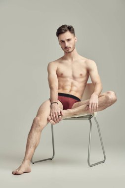 Sandalyenin üzerinde beyaz bir stüdyo arka planına karşı erkek donu içinde çıplak poz veren yakışıklı bir atletin tam boy portresi. İç çamaşırı ve erkek güzelliği..