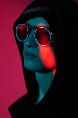 Siyah hırkalı, kapüşonlu ve zarif yuvarlak gözlüklü vahşi bir erkek mankenin portresi. Stüdyo, karışık renkli ışıklandırmayla çekildi. Gözlük stili. 