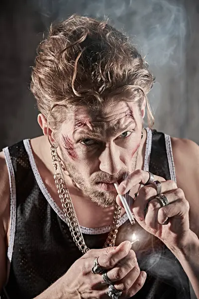 Kötü adam konsepti. Yüzünde morluklar ve yaralar olan, beton duvarlı karanlık bir odada sigara içen sert bir erkek haydutun fotoğrafı. Aksiyon filmi kahramanı.
