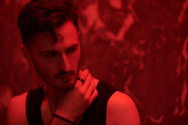 Men's emotions and beauty. Art portrait of a masculine handsome brunette man, who is looking ahead warily and confidently, standing in a dark room with red lighting. Beams of light fall on him.