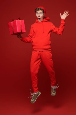 Kırmızı kapüşonlu ve kırmızı şapkalı neşeli bir genç bir çocuk hediye kutusuyla zıplıyor ve büyük gözleri olan şaşkınlıkla bakıyor. Kırmızı bir arkaplanda stüdyo boy portresi. Kutlama, kutlama..