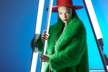 Yüksek moda ve tarz. Parlak makyajlı, sarı saçlı, şık yeşil kürklü ve neon lambaların arasında kırmızı şapkalı çarpıcı bir manken kız. Mavi arkaplanda stüdyo resmi. 