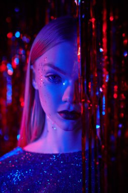 Parlak parlak makyajlı ve parlak bir gece elbisesi olan çekici sarışın bir kız kırmızı folyo perdelerin arasında duruyor. Gece kulübü, disko, neon ışıklar. Parıltı, moda ve tarz. 