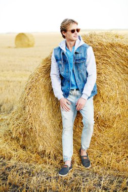 Moda giysiler ve güneş gözlüğü takan mutlu genç adam bir sonbahar tarlasında samanlığın yanında duruyor ve geniş bir şekilde gülümsüyor. Erkek kot giyimi. Gözlük stili.