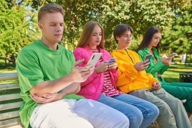 Modern nesil. Parlak gençlik kıyafetleri içinde parktaki bankta ellerinde telefonlarla oturan gençlerin portresi. Gençlik yaşam tarzı. Gençlik yaşı. 