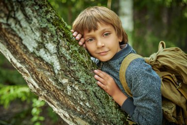 Eko-turizm. Sırt çantalı mutlu bir çocuk yaz ormanında duruyor ve neşeyle bir ağaca sarılıyor. Macera ve seyahat tutkusu.