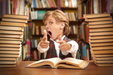 Temiz üniformalı duygusal bir öğrenci, okul kütüphanesinde kameraya korkuyla bakan kitap yığınlarının arasında oturuyor. Modern eğitim ve çocuklar.
