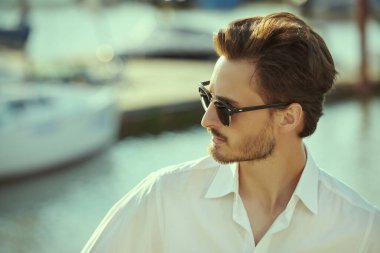 Zarif gömlekli ve siyah güneş gözlüklü yakışıklı bir esmer yat limanında duruyor. Deniz yolculukları. Yaz tatili. Erkek yaşam tarzı. Zarif erkek modası ve tarzı. Boşluğu kopyala.